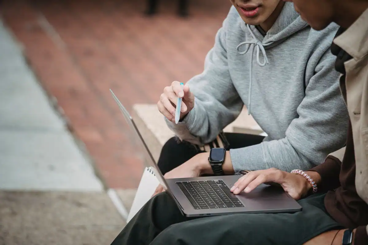Studenți așezați pe o bordură de piatră care compară informații despre facultăți pe laptop