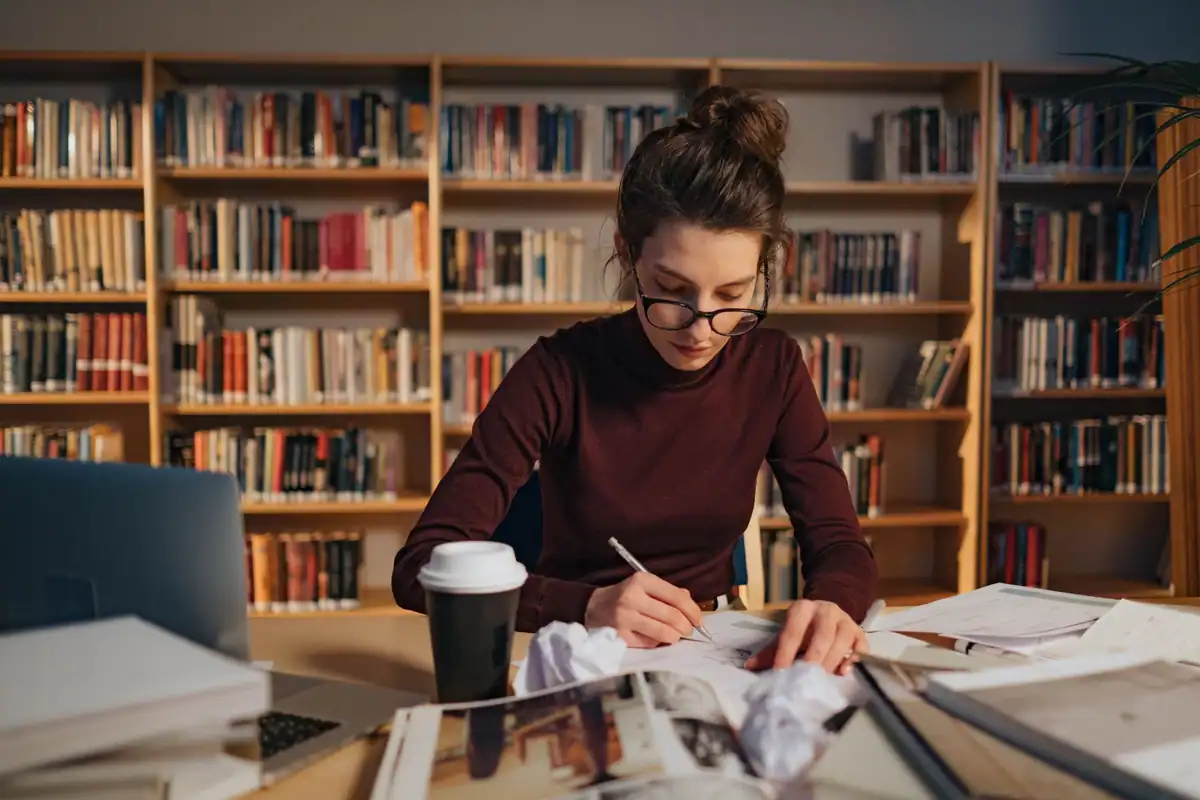 Studentă concentrată care învață într-o bibliotecă, cu rafturi pline de cărți în spate
