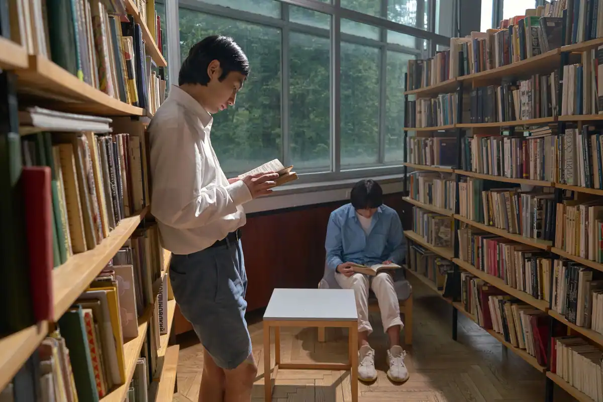 Doi cititori într-o bibliotecă luminoasă, unul în picioare și unul așezat cu cartea