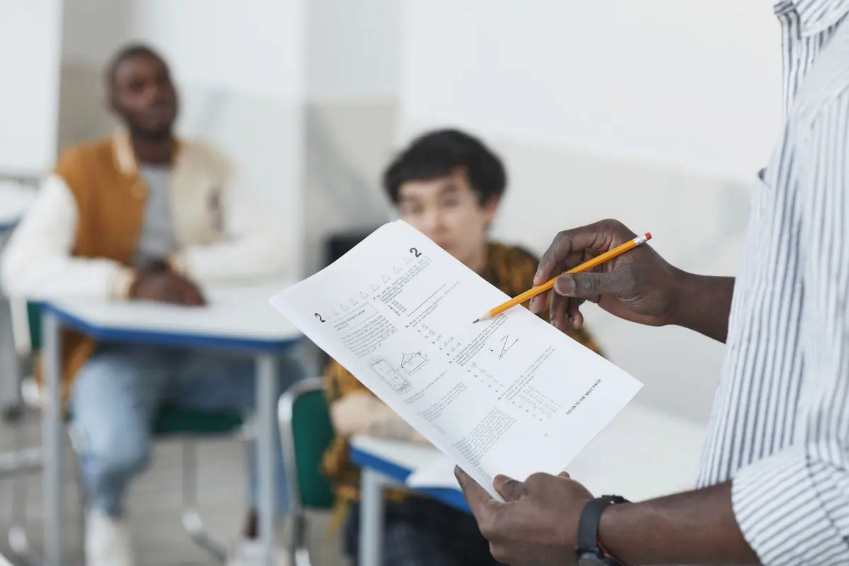 Profesor care verifică foaia de examen a unui elev, în timp ce colegii lucrează