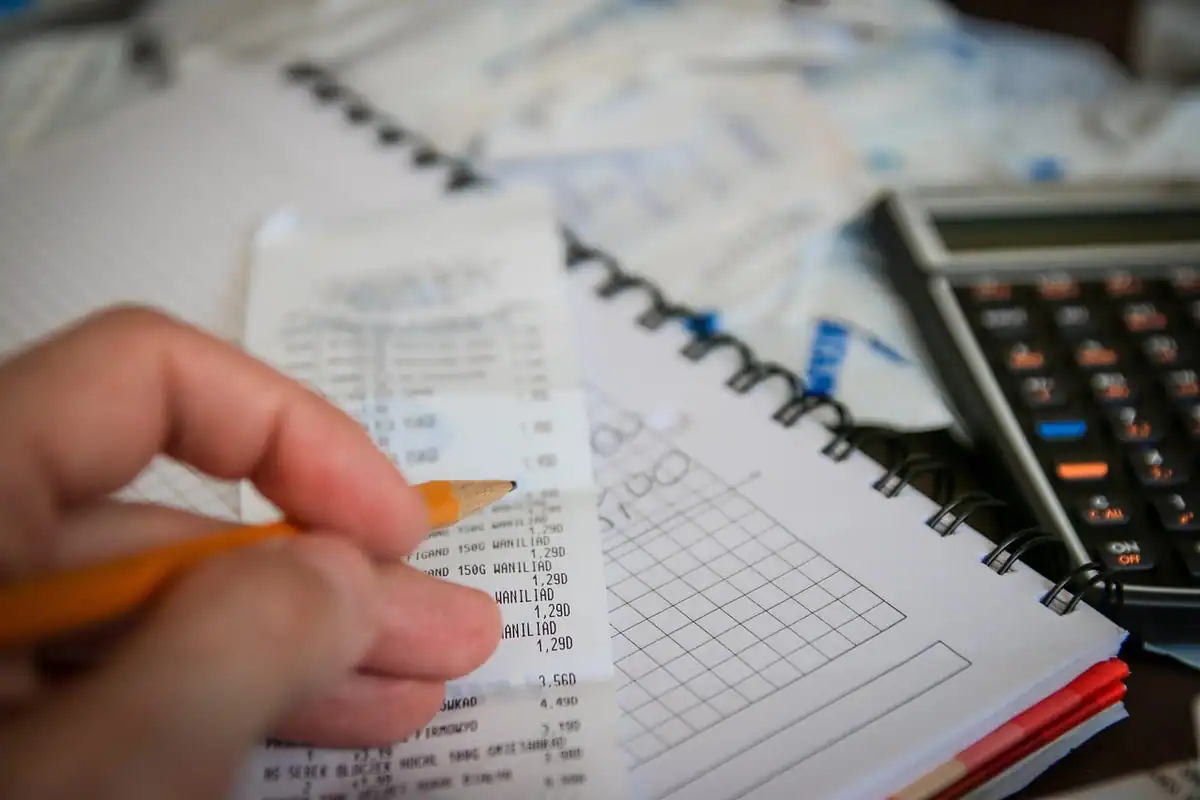 Mână care ține un bon lângă calculator și caiet, socotind cheltuielile de studiu
