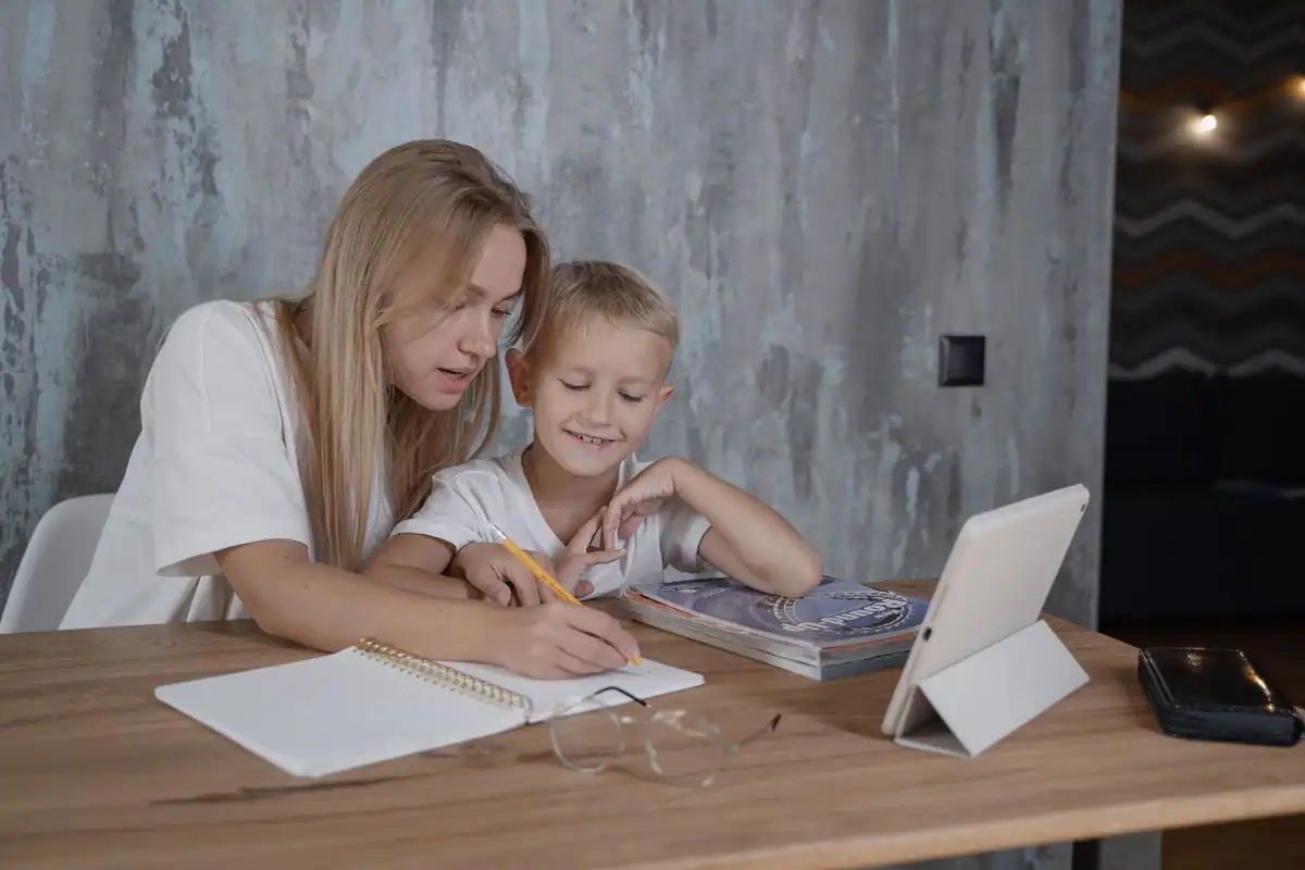 Mamă care își ajută băiatul la teme cu tableta și caietul la masă