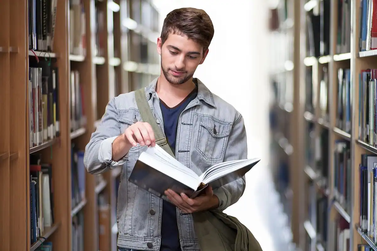 Student cu rucsac care răsfoiește o carte printre rafturile bibliotecii universitare